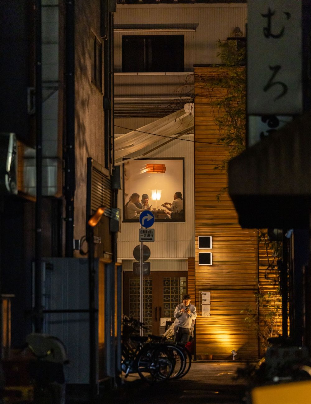 Nighttime street scene in Japan with people dining in a warmly lit restaurant above a narrow alley with parked bicycles.