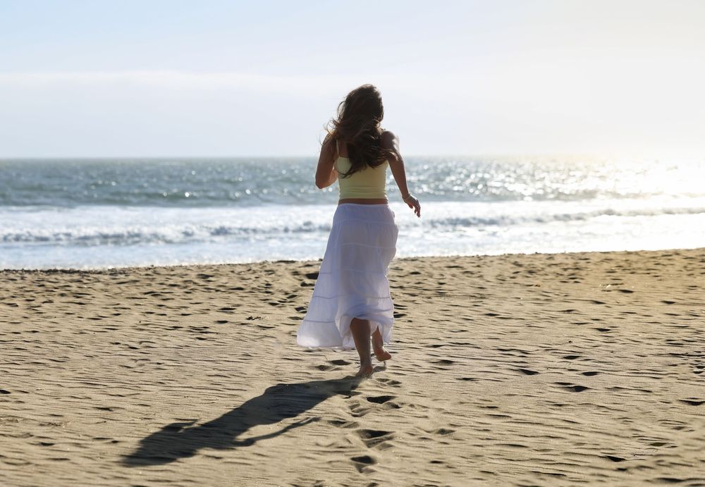 Woman in white skirt running on sandy beach toward ocean waves on a sunny day.