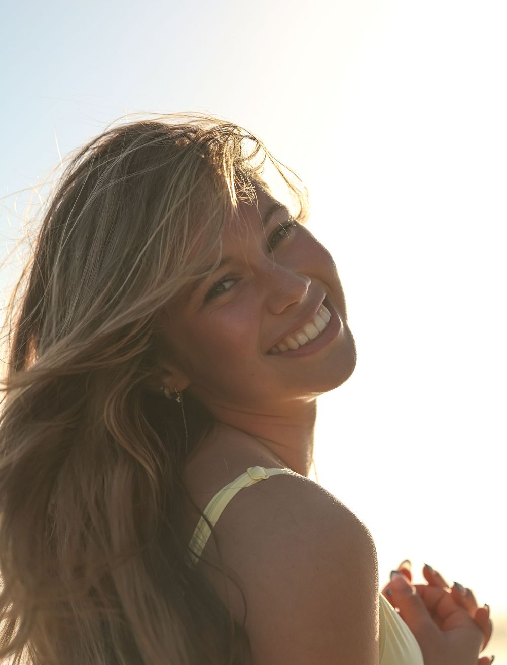 Smiling blonde woman with flowing hair backlit by bright sunlight, wearing a yellow top outdoors.