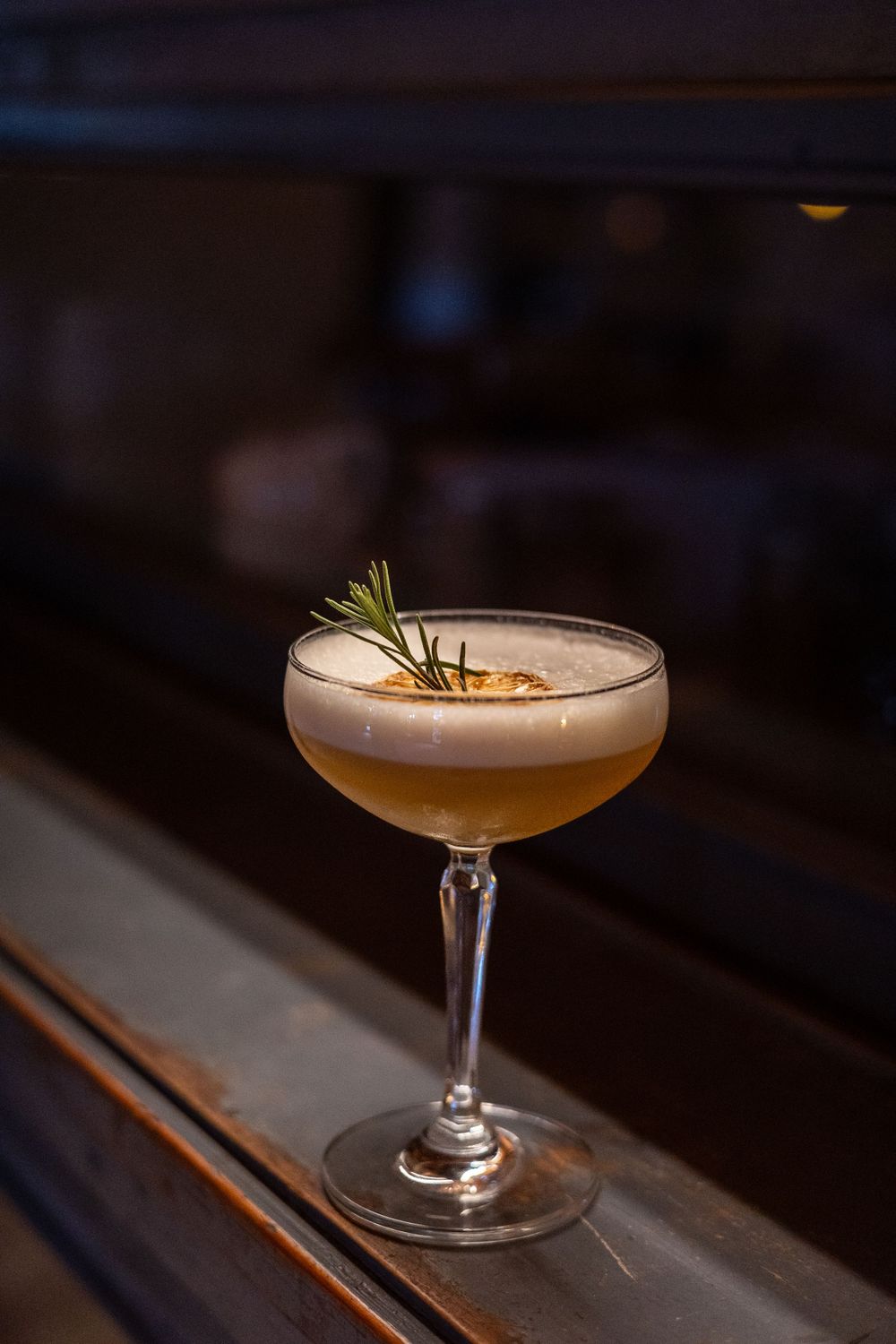 An elegant cocktail in a coupe glass garnished with rosemary and foam sits on a dark wooden surface in moody lighting.