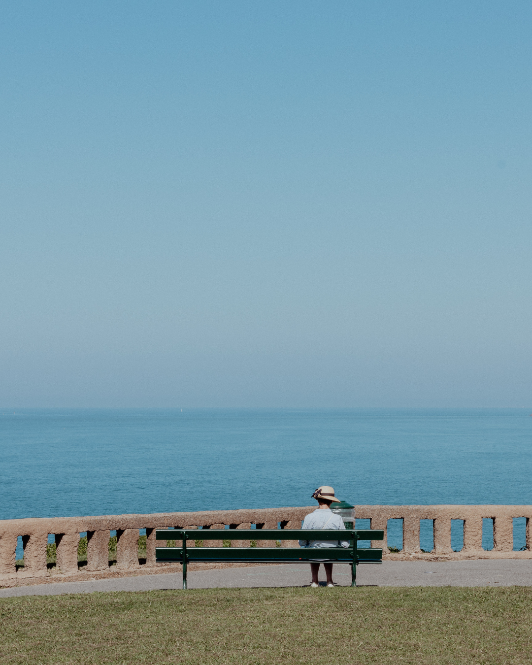 Person sitting alone on bench overlooking ocean view with clear blue sky.
