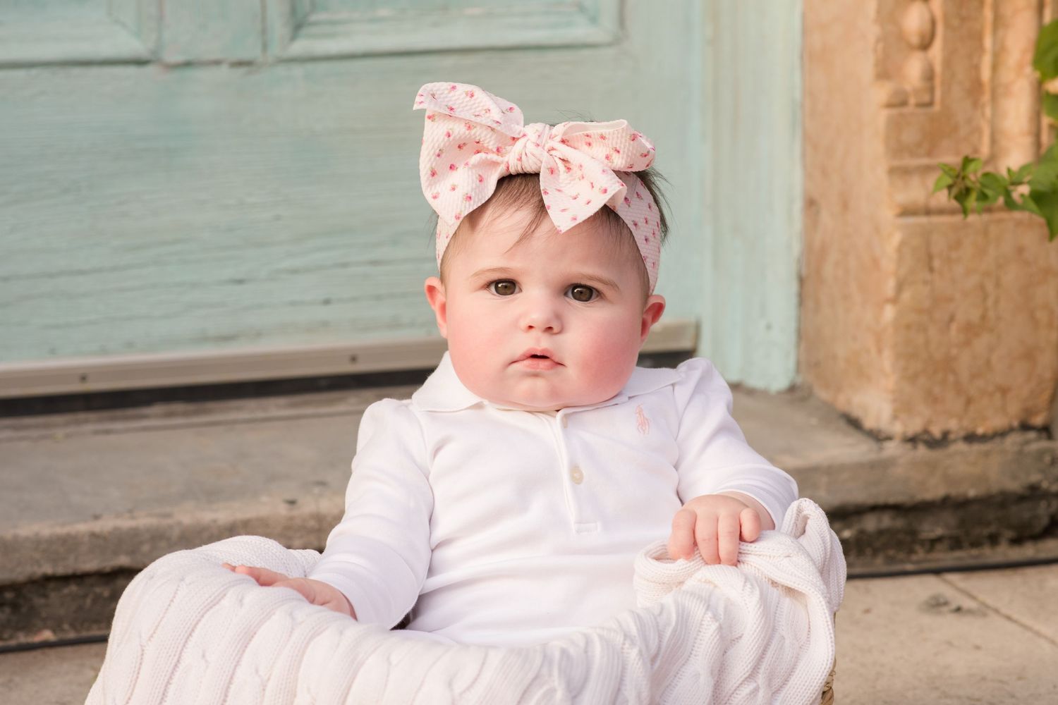 Baby in white dress and bow sits on ornate vintage chair outdoors.