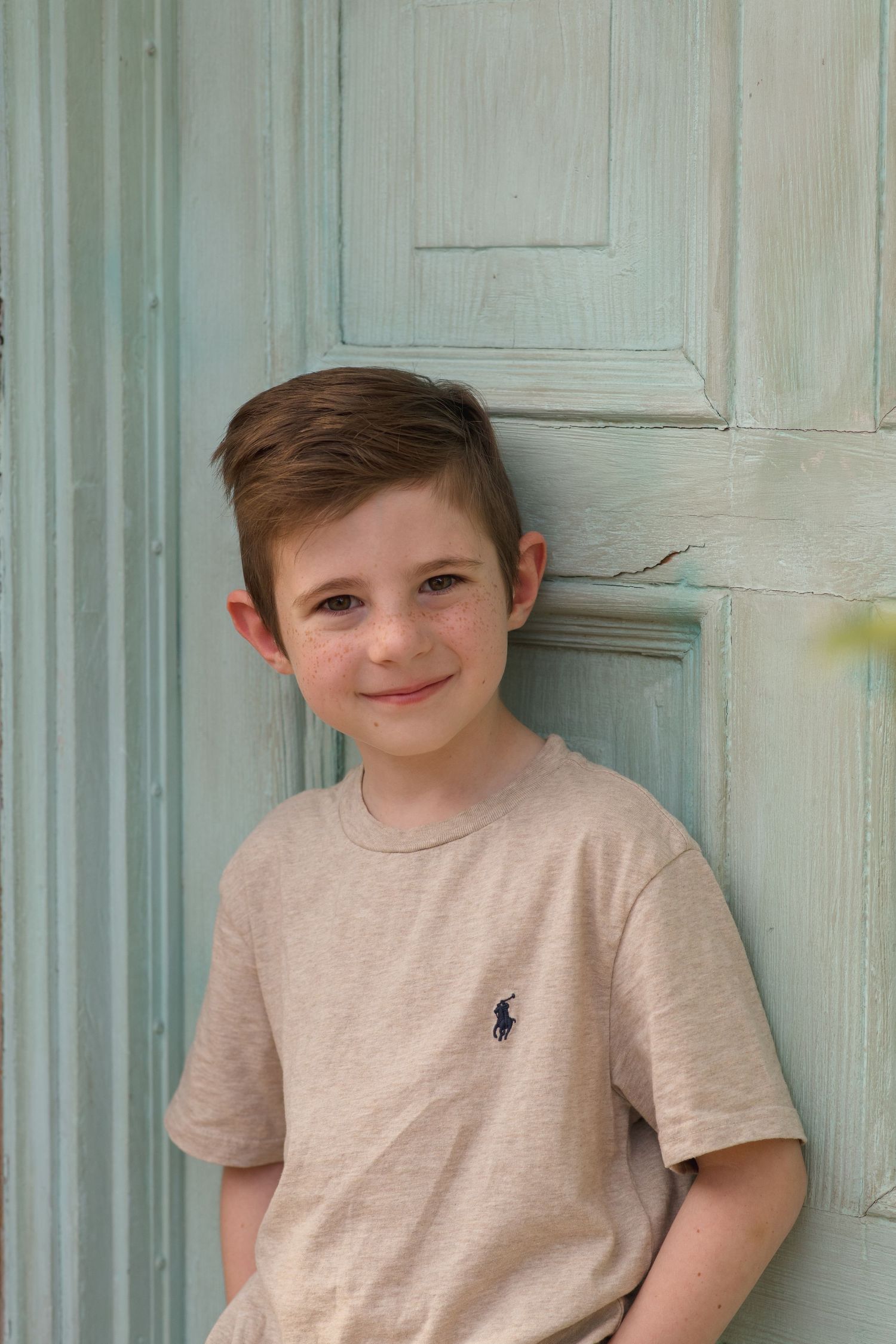 Child in a beige t-shirt with navy logo smiling against a blue wall.