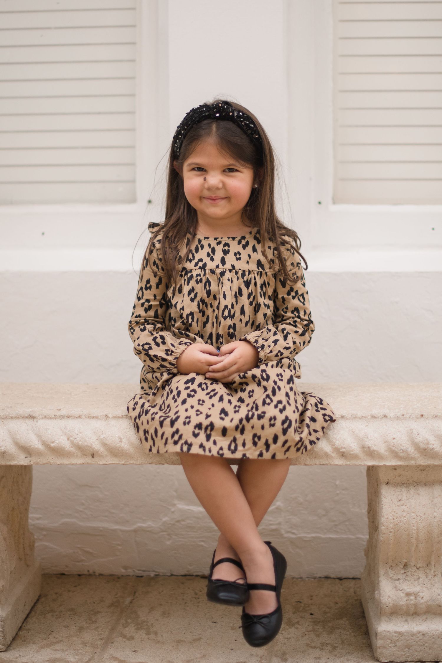Young girl in leopard print dress sitting on stone bench with white shutters behind.
