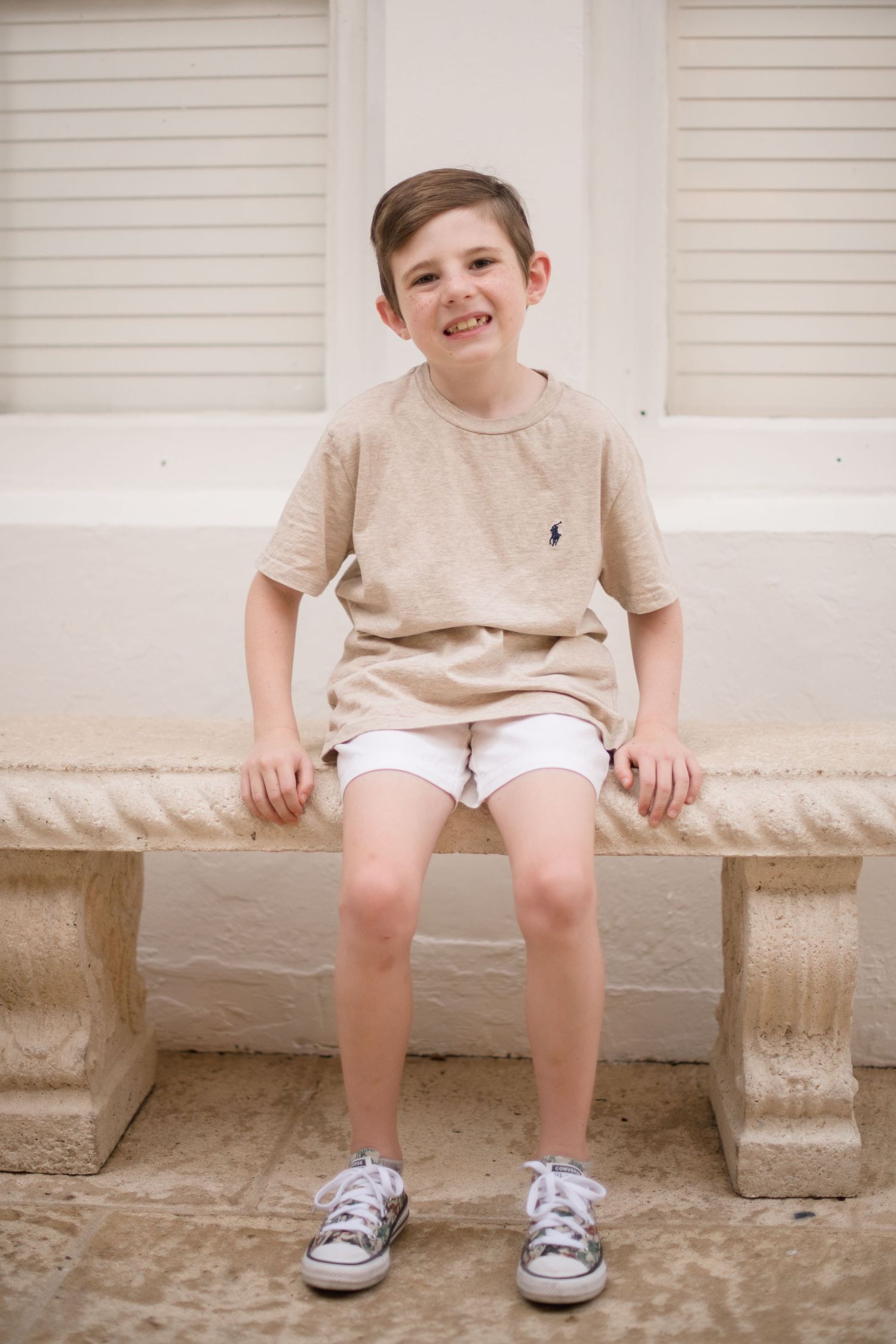 Child in beige polo shirt and white shorts sitting on stone bench.