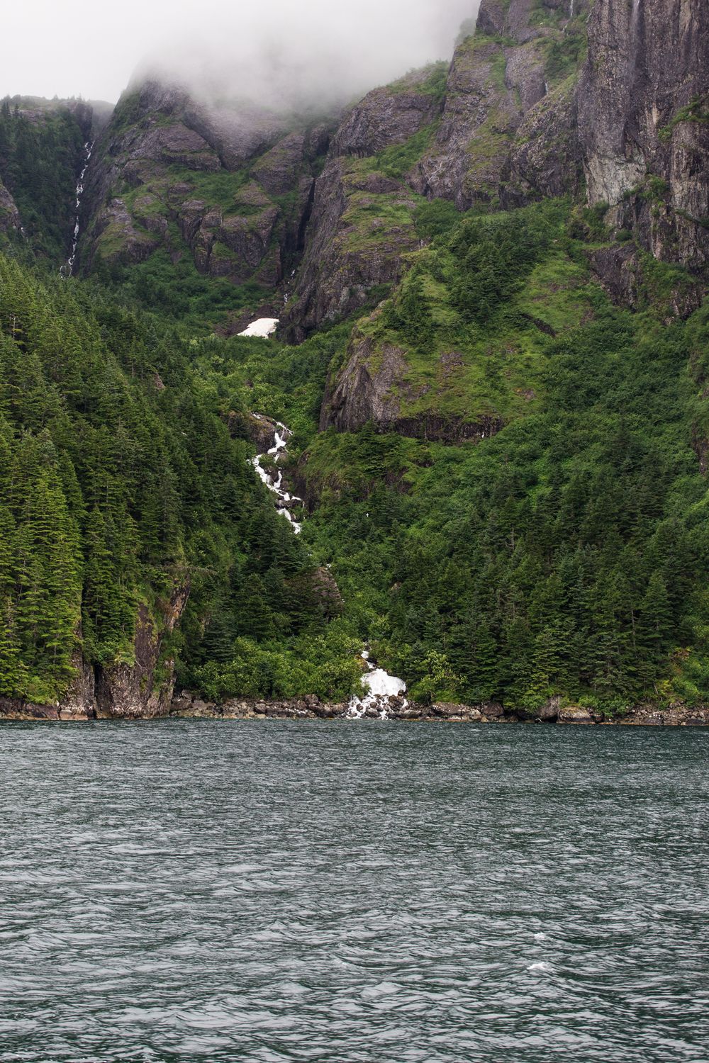 Majestic waterfall cascades down steep rocky cliffs into a fjord surrounded by lush green vegetation.