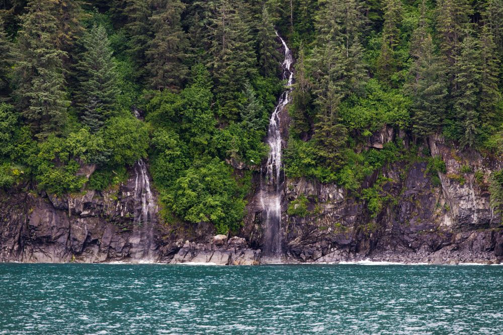 Cascading waterfalls pour down rocky cliffs into turquoise waters along a forested coastline.