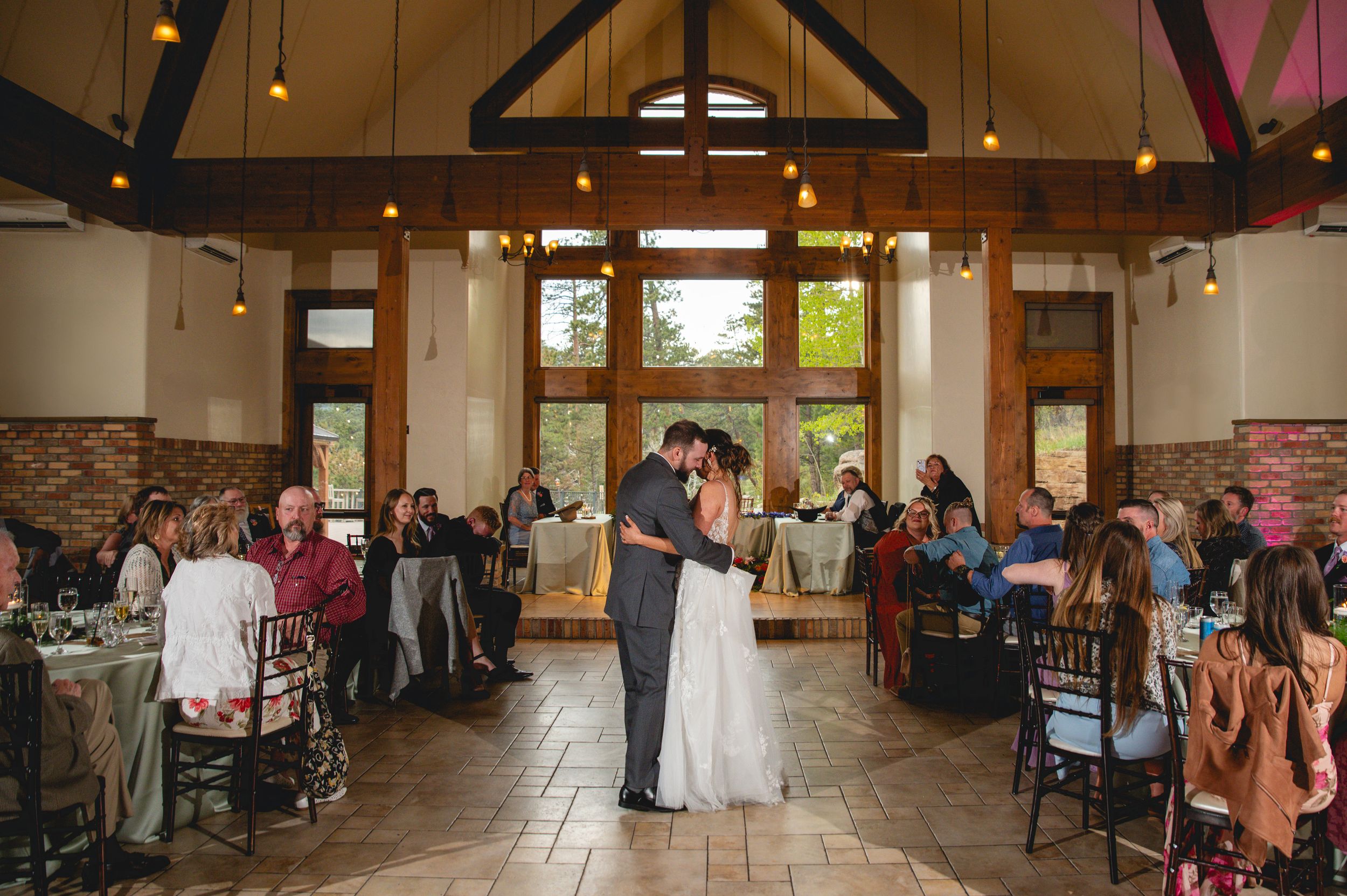 Two people in formal wedding attire stand at a ceremony altar in a rustic indoor venue with wooden beams.
