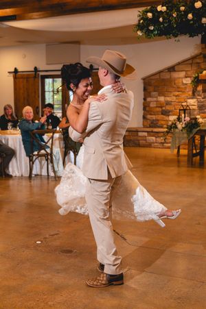 Groom in tan suit and cowboy hat lifts smiling bride in white lace gown during first dance at rustic wedding venue.