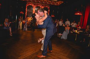 Couple sharing romantic first dance lift at wedding reception on wooden dance floor with guests watching in dimly lit venue.
