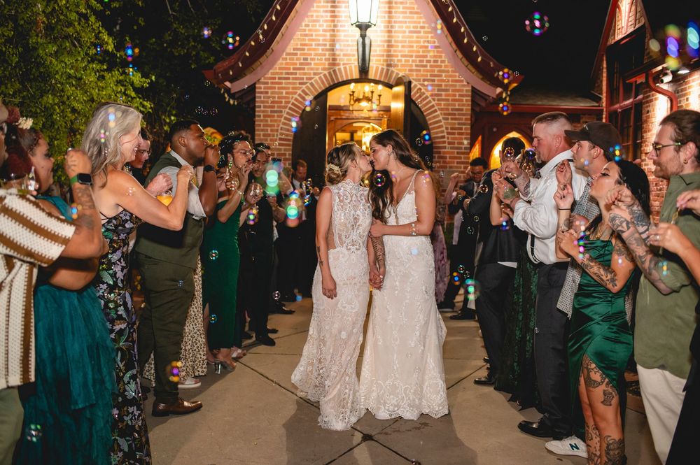 Wedding guests line a walkway outside illuminated brick chapel raising sparklers as newlyweds in white dresses depart.