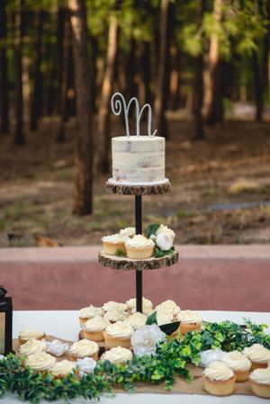A rustic three-tiered wedding dessert display with white cupcakes and a small cake topped with a cursive M in a wooded setting.