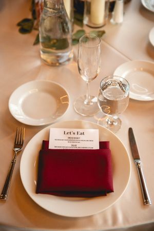 Elegant place setting at wedding reception with burgundy napkin and 'Let's Eat' menu card.