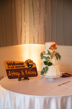 Wedding cake and wooden welcome sign on table with white tablecloth.