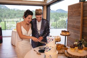 Couple laughing while cutting their multi-tiered wedding cake with mountain views in background.