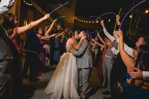 Newlyweds share a kiss under an arch of colorful glow sticks held by cheering wedding guests at a nighttime reception.