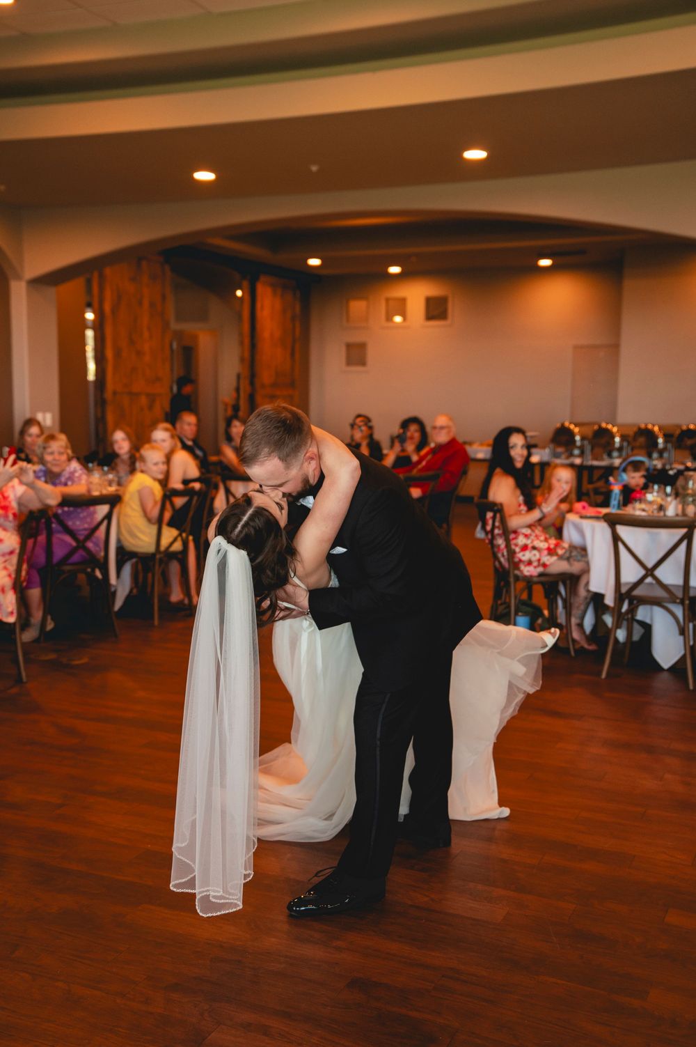 A group of wedding guests dance together on a polished hardwood floor at an indoor reception venue.