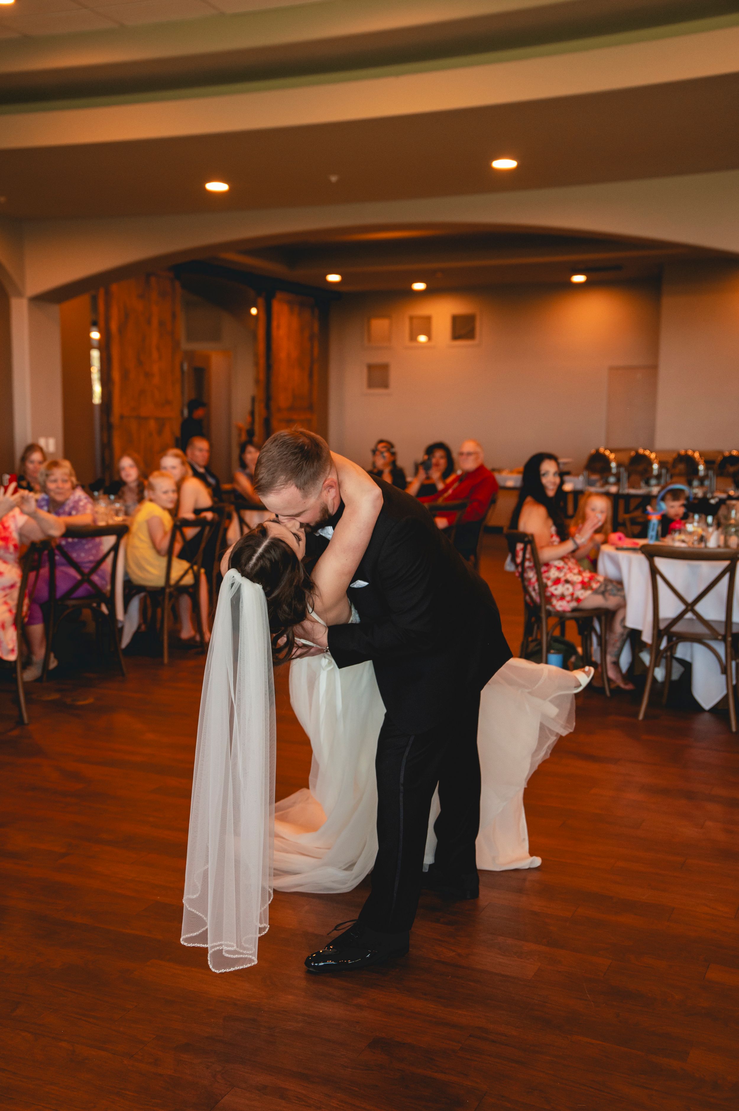 A group of wedding guests dance together on a polished hardwood floor at an indoor reception venue.