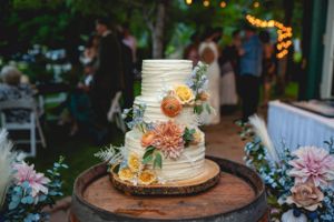 A textured white wedding cake decorated with peach and yellow flowers on a wooden stand at an outdoor reception.