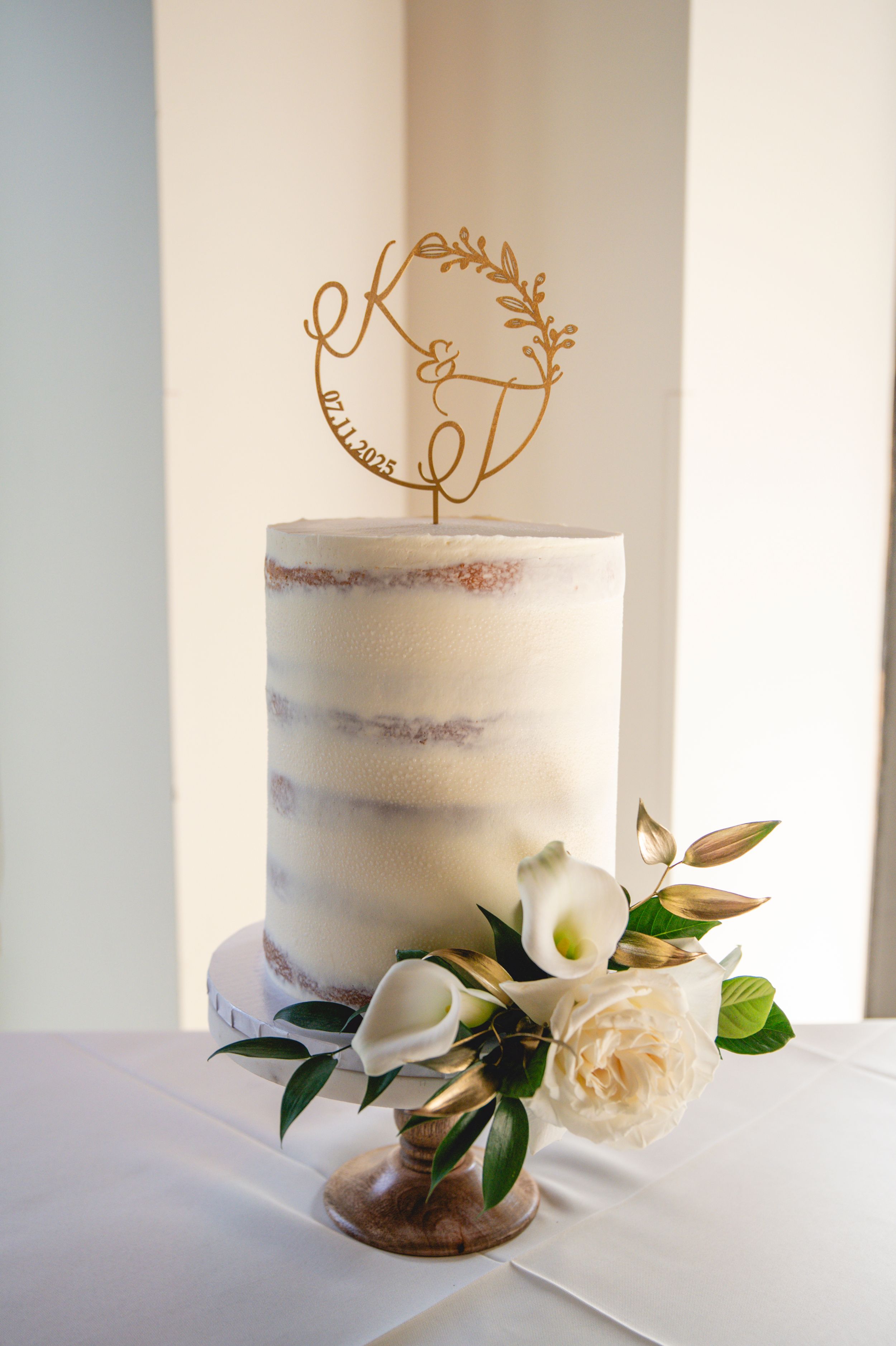 Simple white semi-naked wedding cake decorated with a delicate gold wire topper and fresh green foliage.