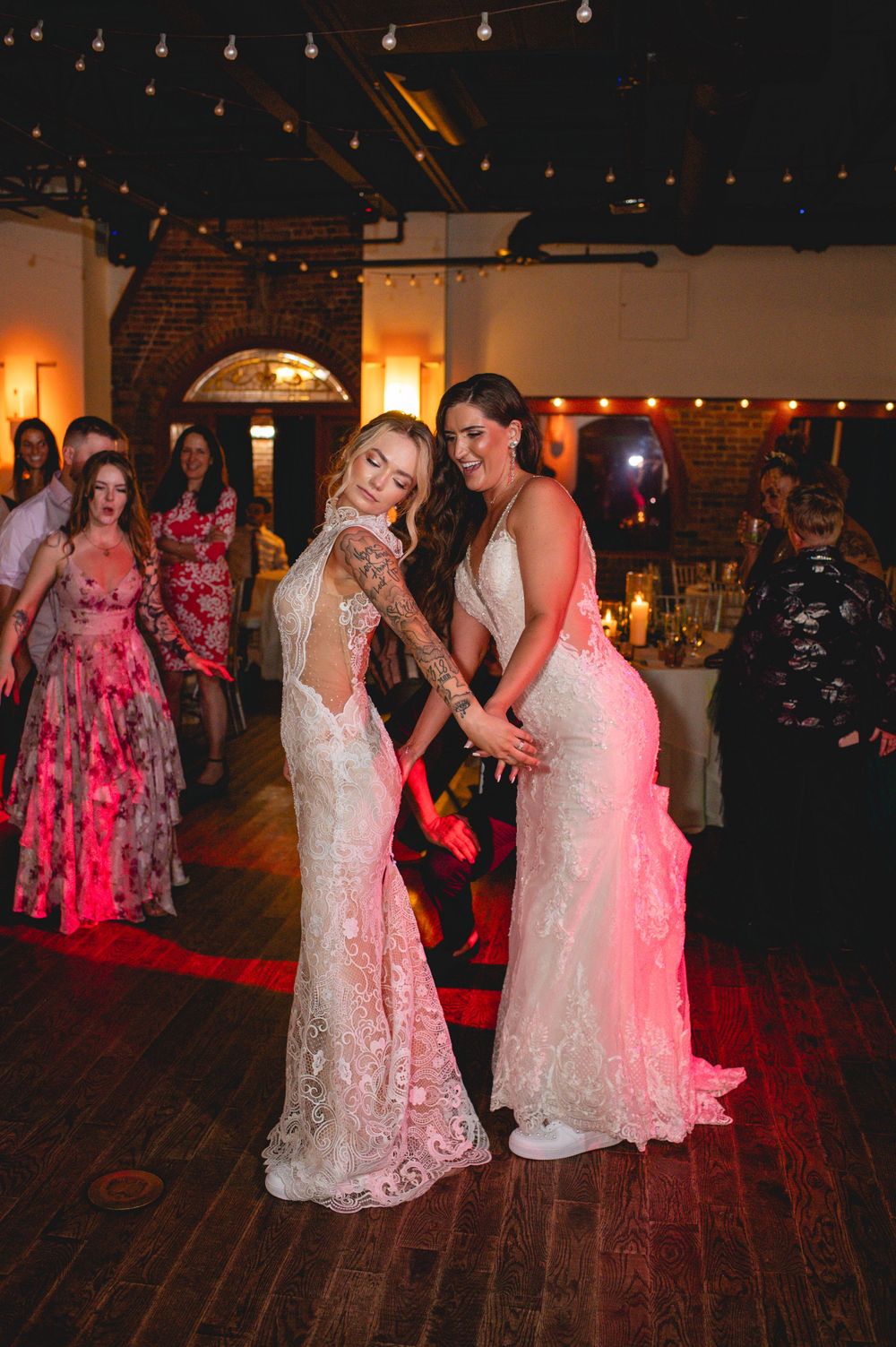 Two women in white lace wedding gowns dance together on a wooden floor under string lights at an evening reception.