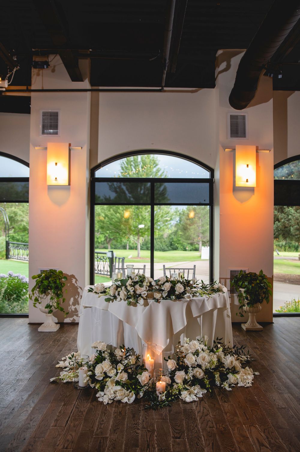 An elegant reception table setting with white florals and candles against arched windows in a venue with warm sconce lighting.