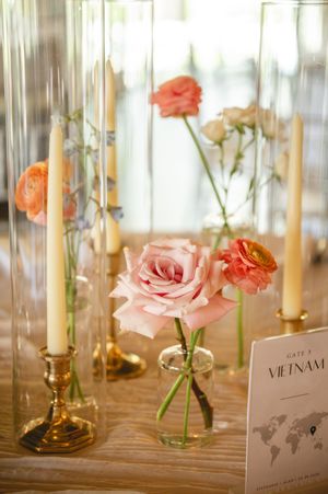 Elegant wedding table centerpiece with pink roses, coral ranunculus, taper candles in gold holders, and a Vietnam gate card.