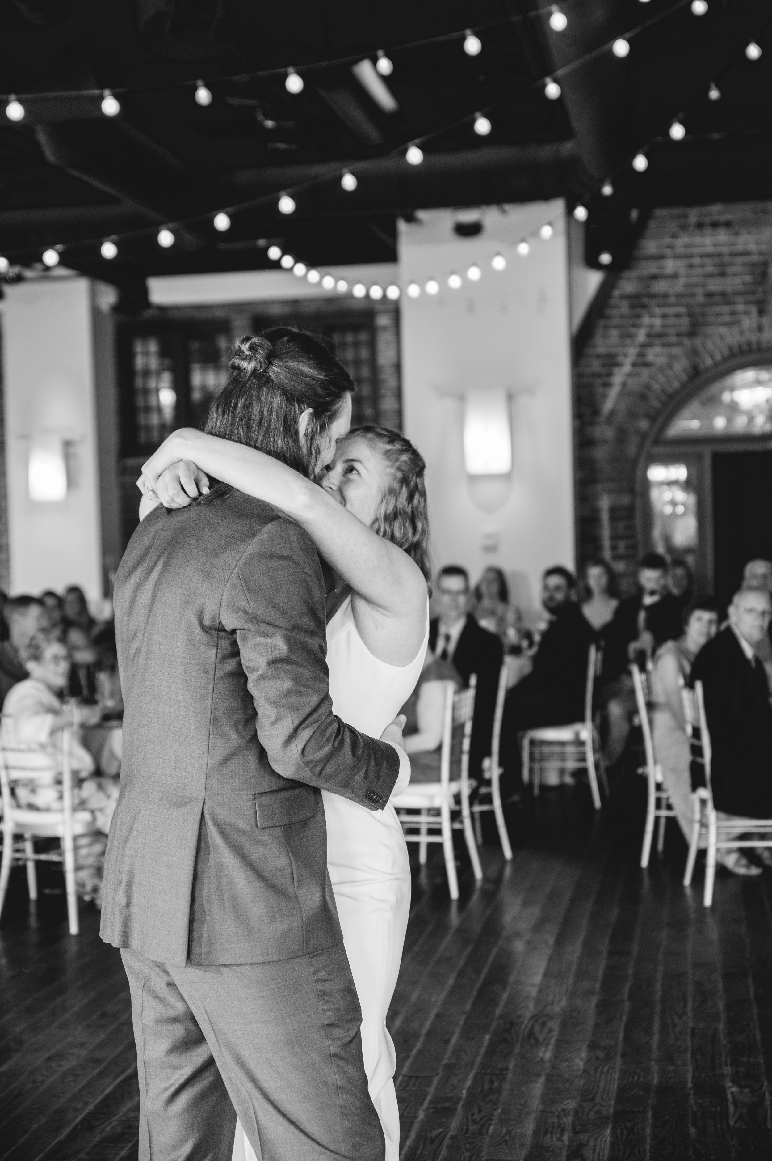 A couple shares their first dance at a wedding reception under string lights with guests watching from white chairs.