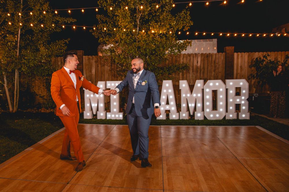 Two people dance on a wooden floor under string lights with illuminated 'MI AMOR' sign in background.