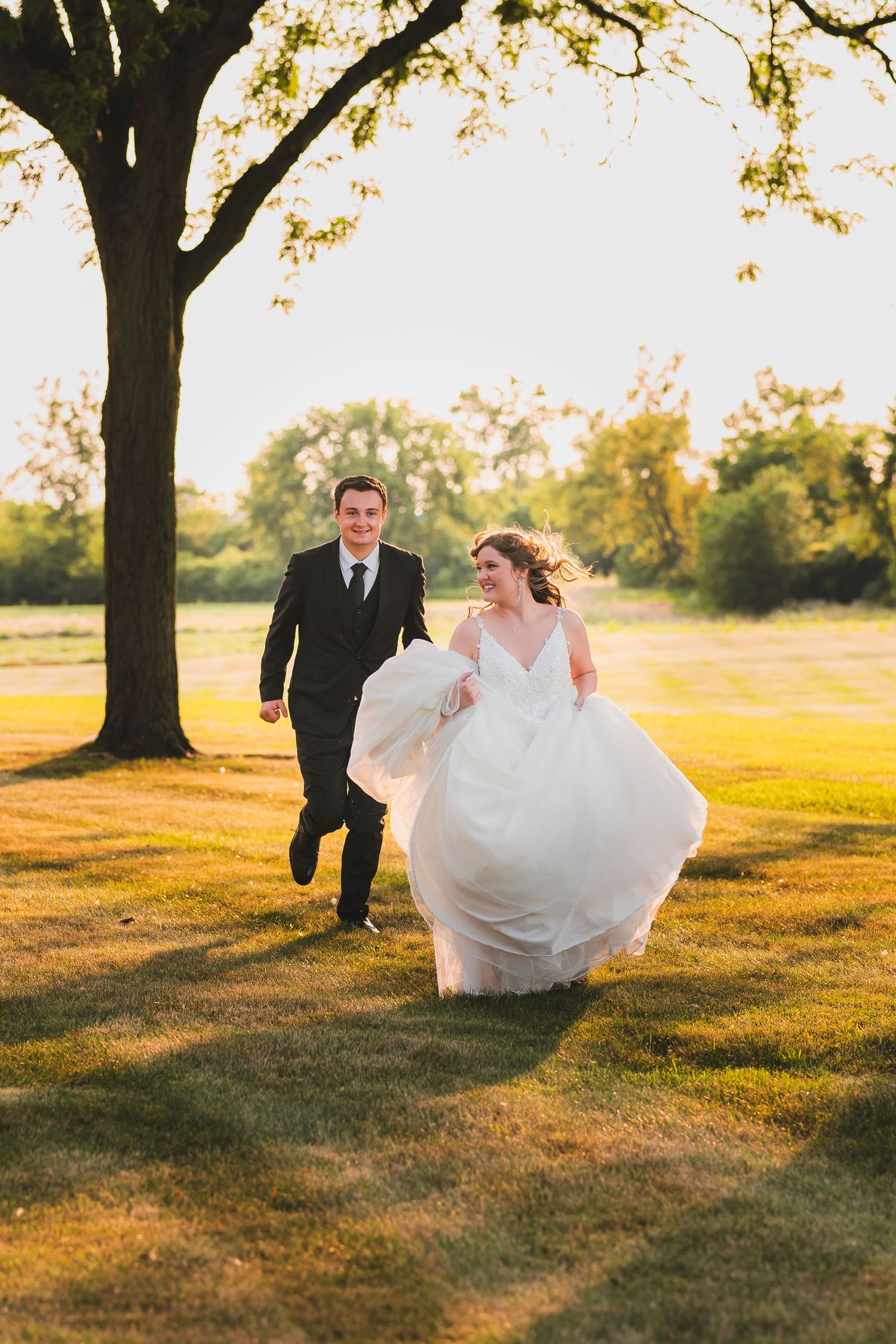 A couple in wedding attire running playfully through a sunlit field under a large tree during golden hour.