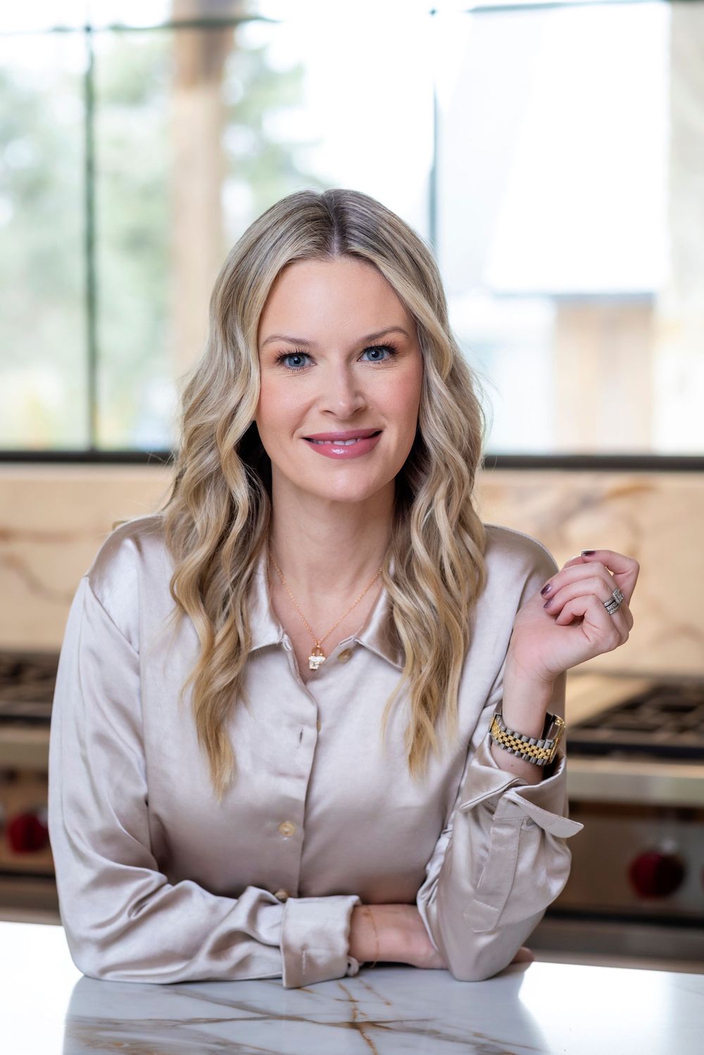 Professional portrait of a person with blonde wavy hair wearing a silk blouse while sitting at a marble counter.