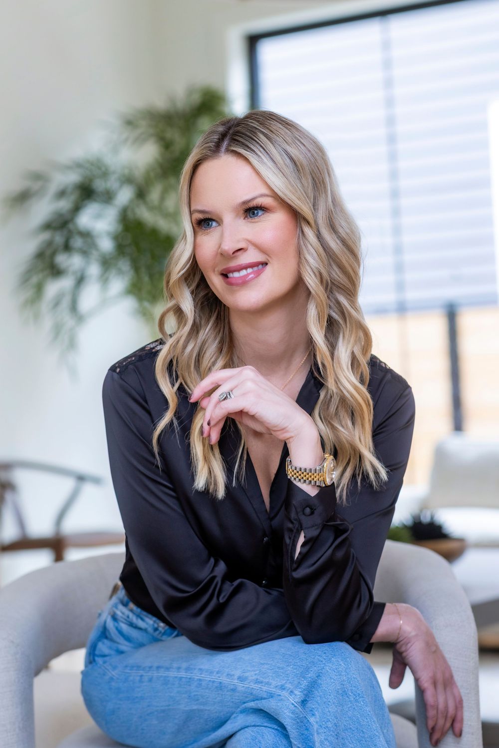 Professional portrait of someone in a black blouse and jeans sitting casually on a chair with indoor plants in background.