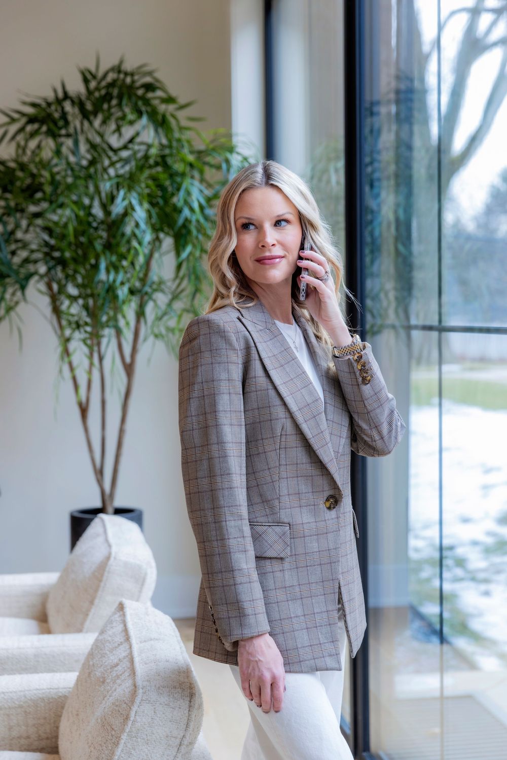 Professional wearing a gray plaid blazer stands in modern office space with large windows and indoor plants.