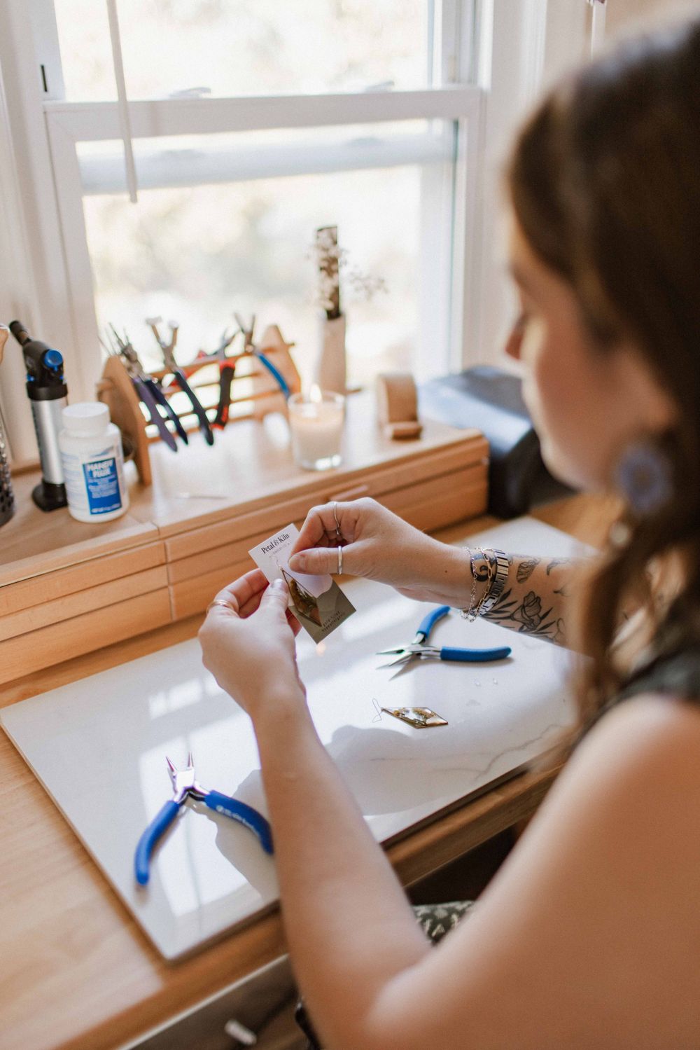 Female jewelry artist working at wooden craft desk near bright window with tools.