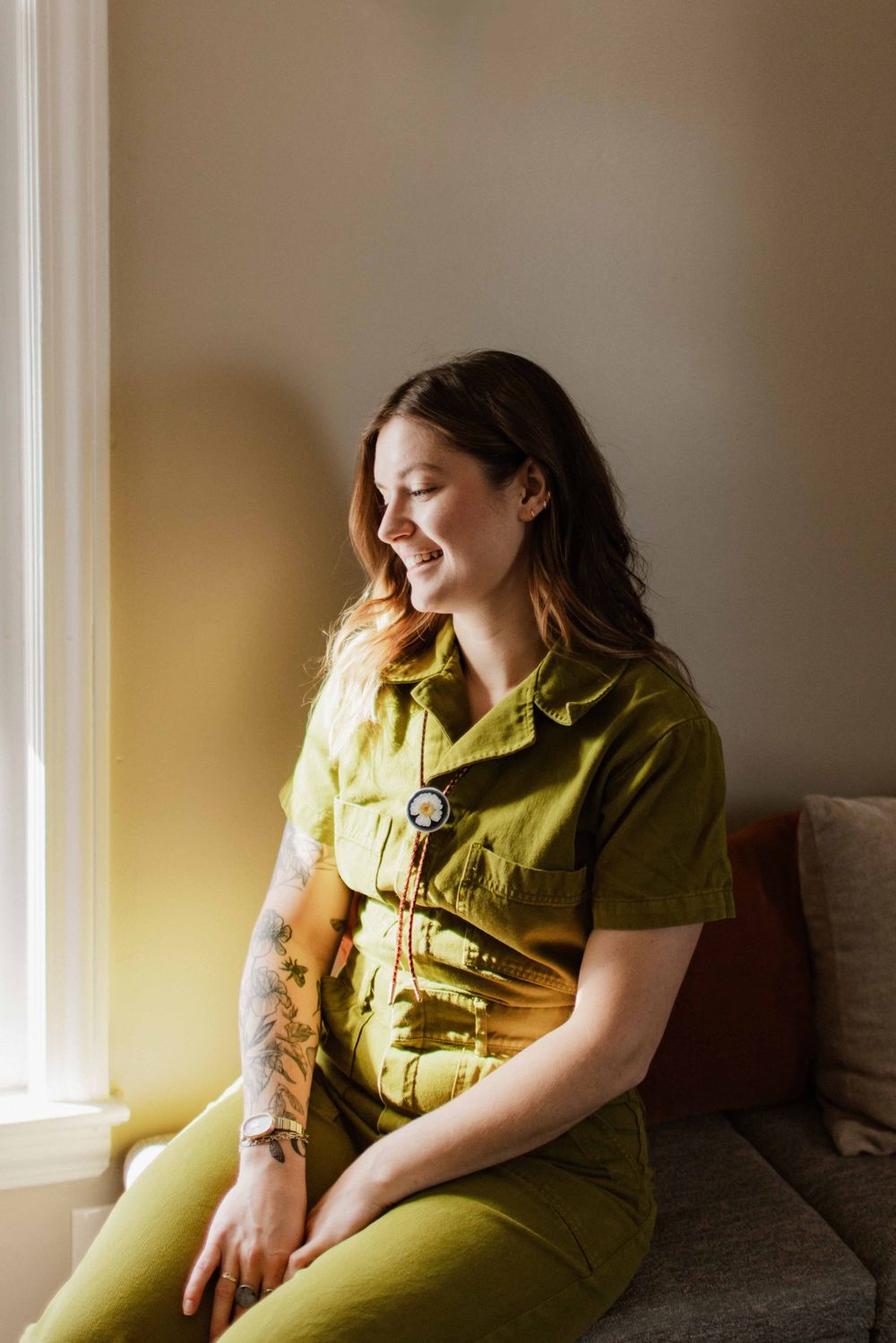 Smiling woman with tattoos wearing olive green jumpsuit sitting by a sunlit window.