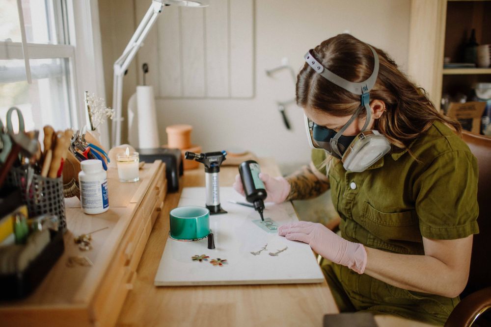 Woman wearing respirator mask and gloves working at a craft jewelry workbench.