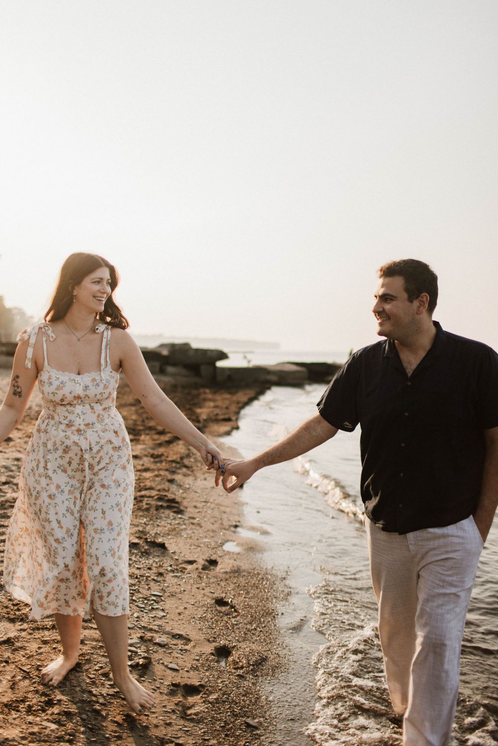 Couple holding hands and walking barefoot along a rocky shoreline at sunset.