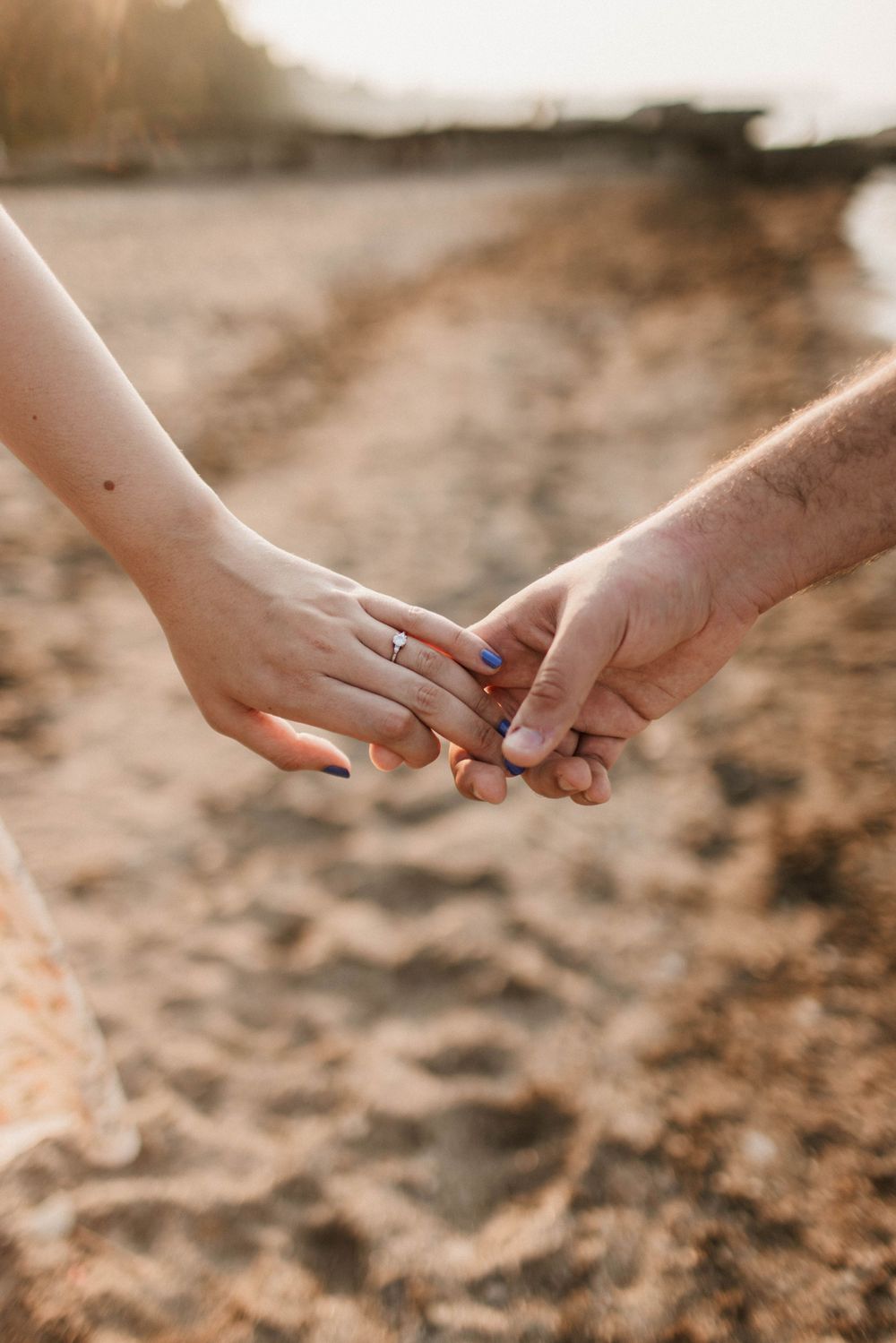 Close-up of couple holding hands on sandy beach with engagement ring visible.