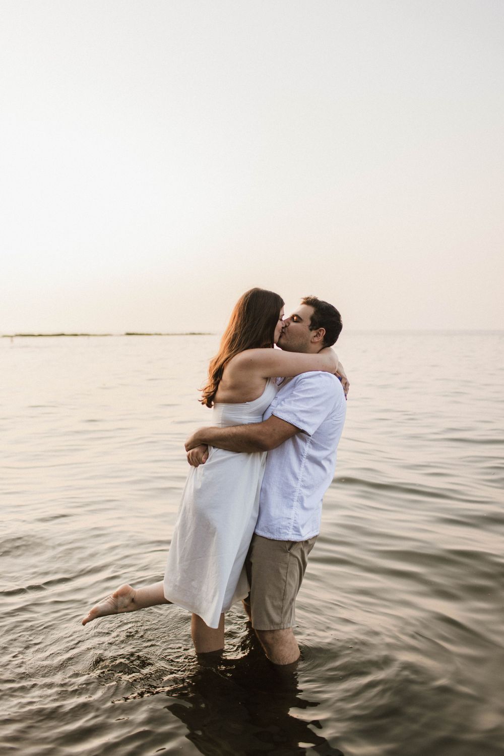 Romantic couple embracing and kissing while standing in shallow ocean water at sunset.