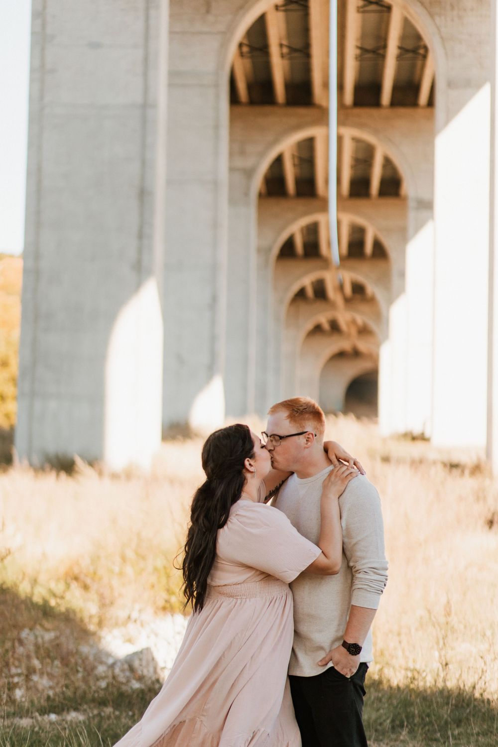 Couple sharing a romantic kiss beneath a large concrete bridge in golden light.