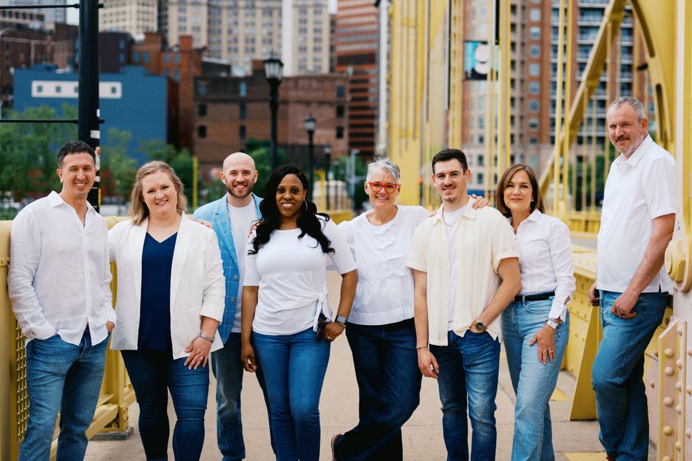A diverse group of professionals in casual attire stands together smiling in an urban setting with city buildings behind them.