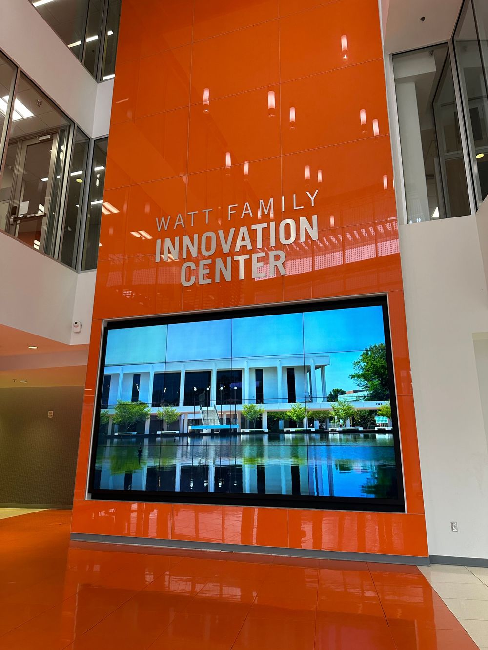 Orange wall display showing Watt Family Innovation Center with digital screen showing modern building reflection in water.