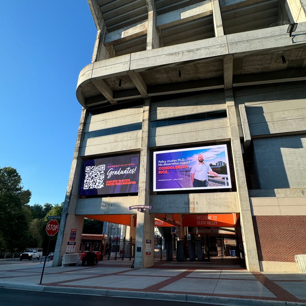 Exterior view of a stadium entrance with digital display screens and concrete architecture against blue sky.