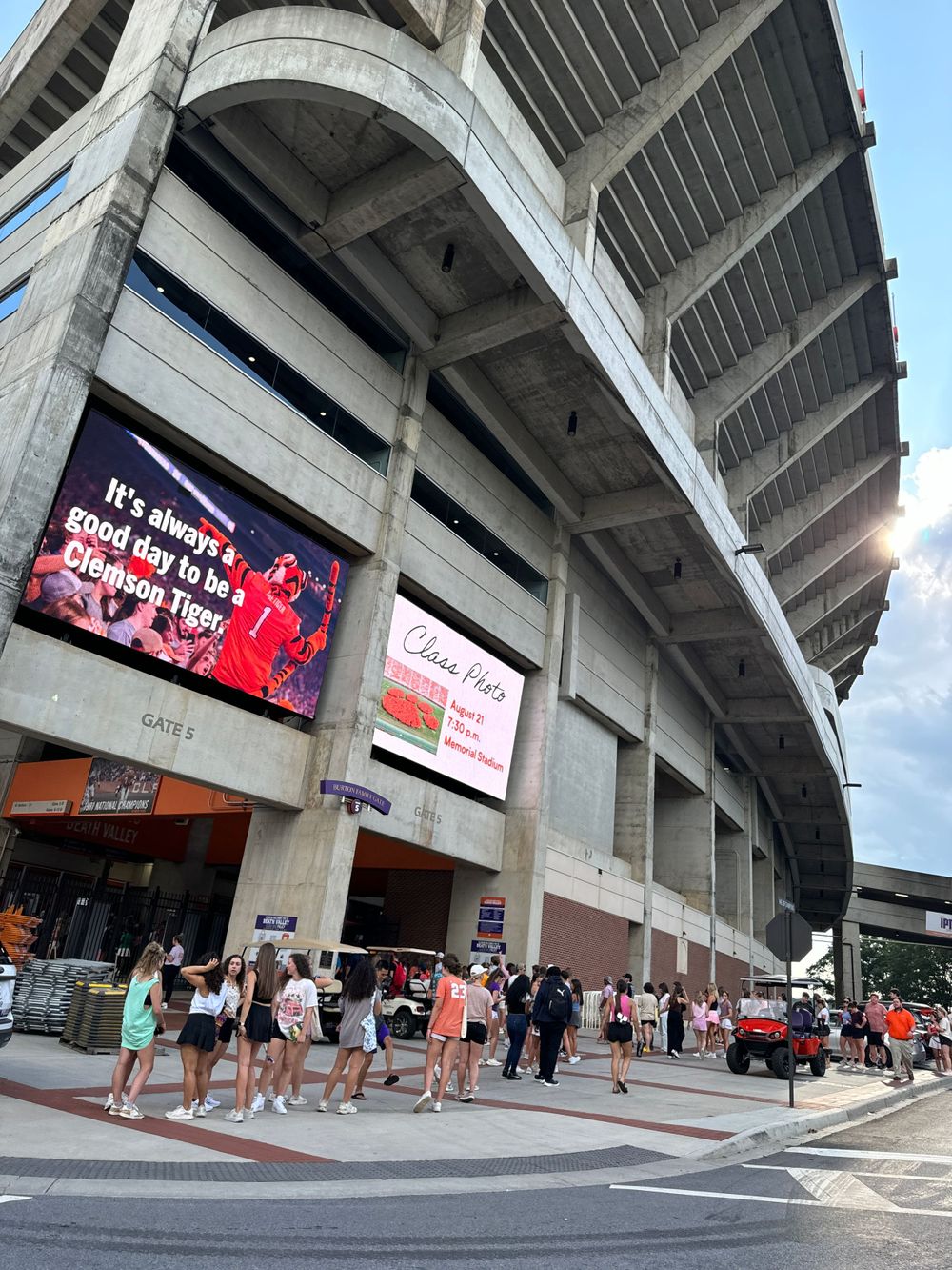 Exterior view of Clemson Memorial Stadium entrance with digital display and fans walking outside.