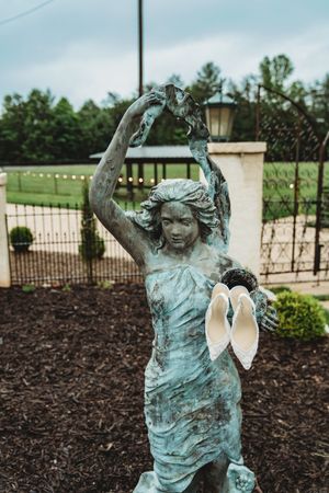 Bronze verdigris female statue holding fabric aloft with white bridal heels hanging from her hand in a garden setting.
