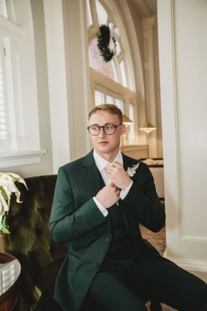 Person in emerald green suit and glasses sitting thoughtfully in elegant room.