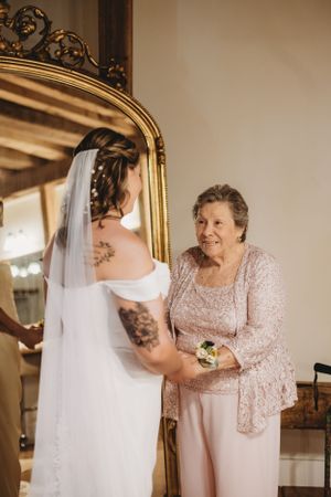 Bride and older woman in pink share a tender moment by ornate gold mirror.