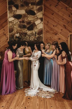 Bride in white gown surrounded by bridesmaids in colorful dresses inside a rustic wood-paneled cabin venue.