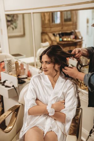 A bride in a white satin robe gets her brunette hair styled by a hairdresser in a elegant salon.