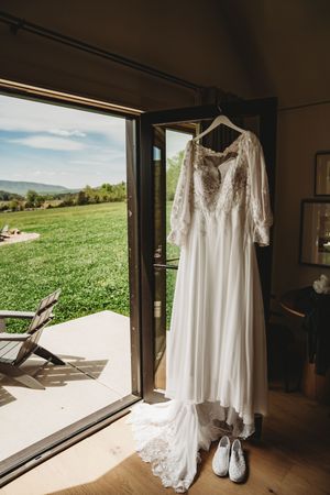 Wedding dress with lace sleeves hanging in doorway overlooking green countryside, with white sneakers below.